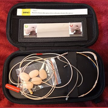 Černý přepravní pouzdro s mikrofonem a příslušením. Uvnitř je béžový headset s červeným konektorem a plastový sáček s několika pěnovými větříky. V ho…