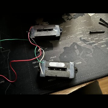 Dvě elektrické kytarové snímače typu humbucker s černým plastovým krytem a viditelnými póly. Na spodní straně jednoho ze snímačů je bílá nálepka s ná…