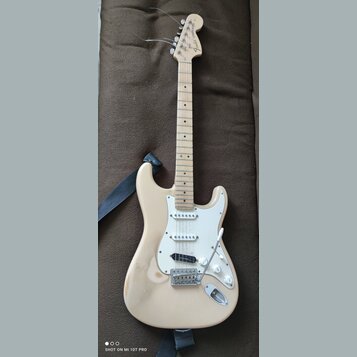 Světle béžová elektrická kytara s bílým pickguardem a javorovým hmatníkem. Na hlavě je nápis Fender STRATOCASTER. Tělo kytary vykazuje známky opotřeb…