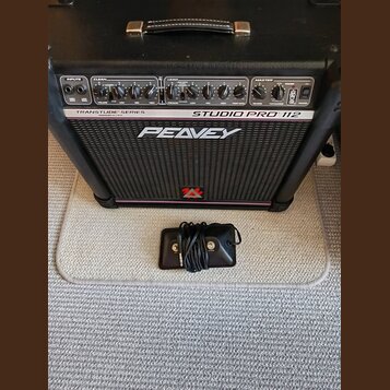 Černý kytarový kombo zesilovač s nápisy TRANSTUBE SERIES, STUDIO PRO 112 a PEAVEY. Na spodní části mřížky je červené logo ve tvaru stylizedého písmen…