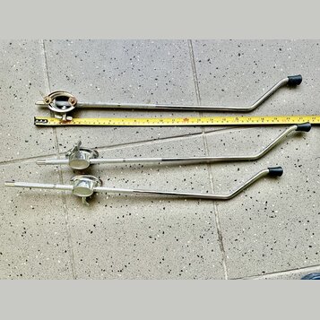 Tři kovové chromované ramena mikrofonních stativů s černými plastovými rukojtěmi. Vedle horního ramene je položen žlutý metr. Na kovových površích js…