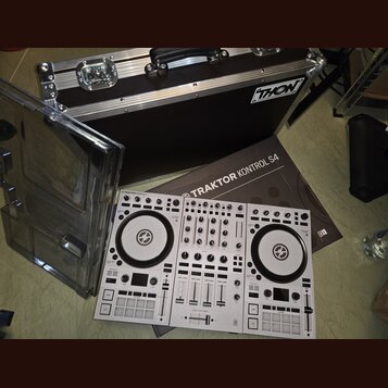 Bílý DJ kontrolér s nápisem TRAKTOR KONTROL S4. Vedle něj je položen hnědý karton s nápisem TRAKTOR KONTROL S4 a v pozadí černý transportní kufr s lo…