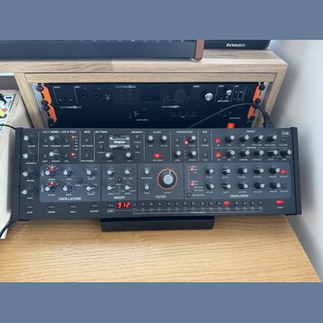 Černý analogový synthesizér s velkým množstvím ovládacích knoflíků a přepínačů. Na panelu jsou vidět nápisy OSCILLATORS, MIXER, FILTER a ENVELOPES. V…