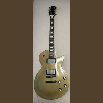 Zlatá elektrická kytara s dvěma humbuckery a čtyřmi ovládacími knoflíky. Na hlavě nástroje není žádný viditelný nápis ani logo. Povrch těla vykazuje…