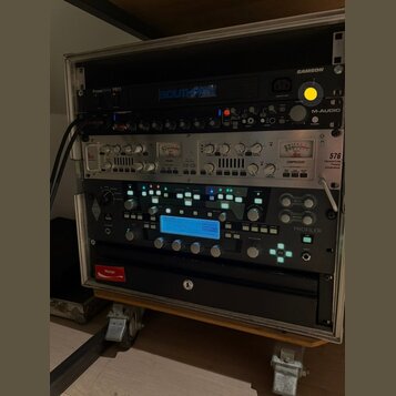 Stojan s rackovým vybavením v hliníkovém kufru na kolečkách. Jsou zde zařízení s nápisy SAMSON, PowerBrite PB11, M-AUDIO a PROFILER. Střední zařízení…
