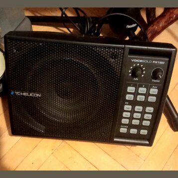Černý kompaktní reproduktor s kovovou mřížkou. Na přední straně je nápis VOICESOLO FX150 a logo TC-HELICON. Ovládací panel obsahuje dva otočné knoflí…