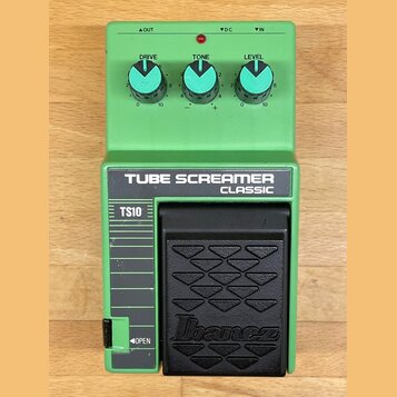 Zelený kytarový efekt s nápisy TUBE SCREAMER CLASSIC a TS10. Na horní části jsou tři modré otočné ovladače s popisky DRIVE, TONE a LEVEL. V dolní čás…