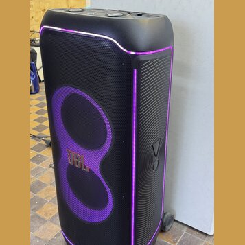 Černý party reproduktor s fialovým LED podsvícením. Na přední mřížce je umístěno zlaté logo JBL. Povrch je kombinací tkaniny a plastu s žebrovaným vz…