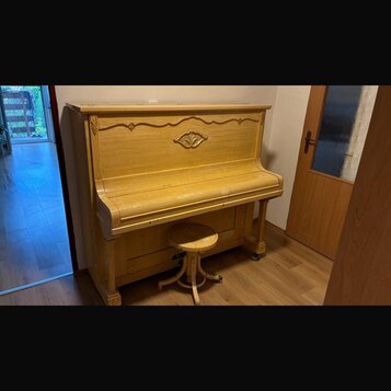 Světle hnědé dřevěné piano s ozdobným vyřezáváním v horní části. Před nástrojem stojí jednoduchá dřevěná taburetka na čtyřnohém podstavci. Povrch pia…