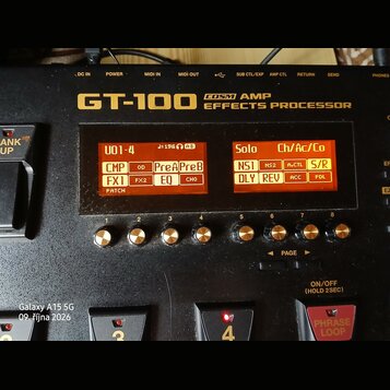Černý efektový procesor s nápisem GT-100 COSM AMP EFFECTS PROCESSOR. Zařízení má dvě oranžové displeje s texty U01-4, Solo a dalšími zkratkami efektů…
