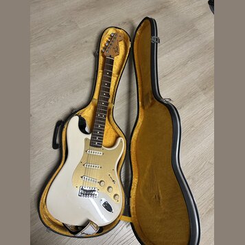 Bílá elektrická kytara s tvarem Stratocaster v černém kufru s žlutým vnitřkem. Na hlavě hředu je viditelné logo Fender. Kytara má tři snímače, zlatý…