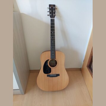 Akustická kytara s přírodním dřevem, černým hmatníkem a černým plektrem. Na hlavě je viditelné logo SIGMA Guitars. Nástroj vykazuje v popředí čistý p…