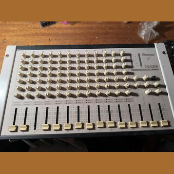 Bíle provedený mixovací pult s nápisem Ibanez RM22 a označením CHANNEL STEREO MIXER. Zařízení obsahuje 12 kanálů s řadou bílých otočných knoflíků a 1…