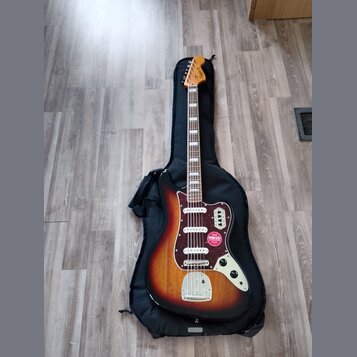 Elektrická kytara s tělem v barvě sunburst a bílým hmatníkem, položená na černém softcasu. Na hlavě je viditelné logo Squier a na těle se nachází čer…