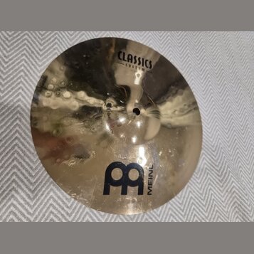 Zlatý kovový činel s viditelným broušením povrchu. Na číneli jsou černé nápisy CLASSICS CUSTOM a MEINL spolu s logem ve tvaru dvou propojených písmen…
