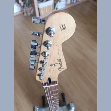 Záběr na hlavu elektrické kytary z přírodního dřeva s chromovanými ladičkami. Na zadní straně jsou nápisy Fender, STRATOCASTER® a ORIGINAL Contour Bo…