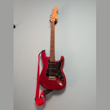 červená elektrická kytara s černým pickguardem a dřevěným hřidnicí. Na hlavě je viditelné logo Fender. Kytara má připojený červený popruh a visí na n…