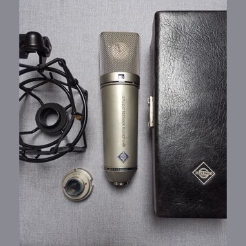 Stříbrný kondenzátorový mikrofon s kovovou síťovkou a nápisem STUDIOTECHNIK RUNDFUNK a MICROPHONE. Na těle je viditelné modré logo ve tvaru kosočtver…