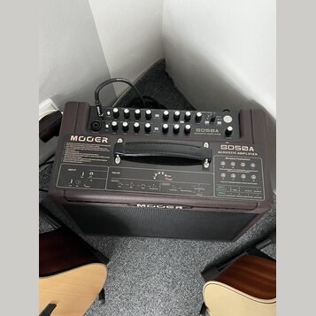 Hnědý akustický zesilovač s nápisem MOOGER a označením modelu SDSØA ACOUSTIC AMPLIFIER. Horní panel obsahuje ovládací knoflíky, přepínače a vstupy, z…
