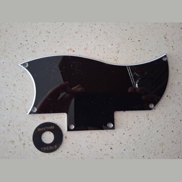 Černý plastový plech ochranný (pickguard) s vyleštěným stříbrným okrajem a otvory pro šrouby. Vedle něj je umístěn malý černý kruhový přepínač s bílý…