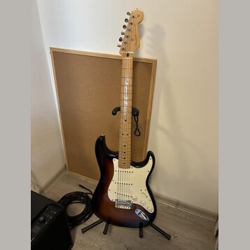 Elektrická kytara s tłem v barvě sunburst a bílým pickguardem. Na hlavě hředu je viditelné logo Fender. Kytara stojí na černém stojanu před korkovou…