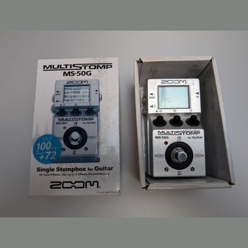 Fotografie zobrazuje stříbrný efektový pedál v krabici vedle původního obalu. Na zařízení i na krabici je vidět logo ZOOM a nápis MULTISTOMP MS-50G f…