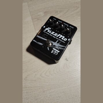 Černý kytarový efekt s bílým nápisem FuzzMo a podtitulem Fuzz Mode Ultra High Gain Distortion. Na spodní části je logo EBS. Zařízení má čtyři ovládac…