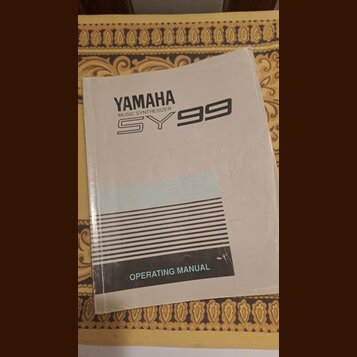 Bílá tištěná příručka s nápisy YAMAHA, MUSIC SYNTHESIZER a SY99. Ve spodní části je černý pruh s textem OPERATING MANUAL. Papír vykazuje známky opotř…