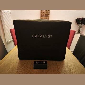 Černý textilní kryt na hudební zesilovač s bílým nápisem CATALYST uprostřed. Před krytem leží černý stupňovací spínací pedál s nápisy CATALYST, CHANN…