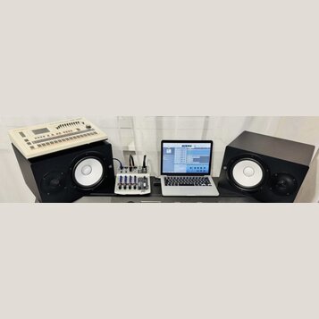 Fotografie zachicuje pracovní stůl s hudební technikou. Vlevo je bílá rytmická sekvenční jednotka s nápisem Roland TR-707, umístěná na černém studiov…