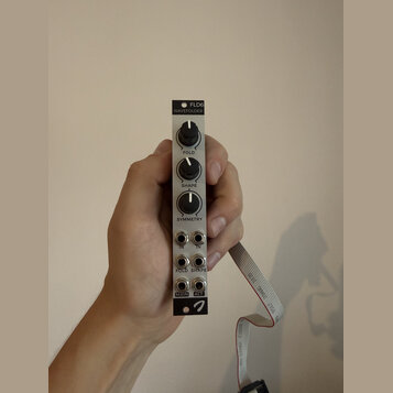 Ruka drží modulární syntetizátor v bílém provedení s nápisy FLD6 a WAVEFOLDER. Zařízení má tři černé otočné ovířkovací knoflíky s popisky FOLD, SHAPE…