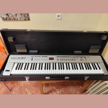 Stříbrný MIDI kontrolér Roland A-37 v černém transportním kufru s polstrováním. Na levé straně je nápis Roland A-37 MIDI KEYBOARD CONTROLLER a vprost…