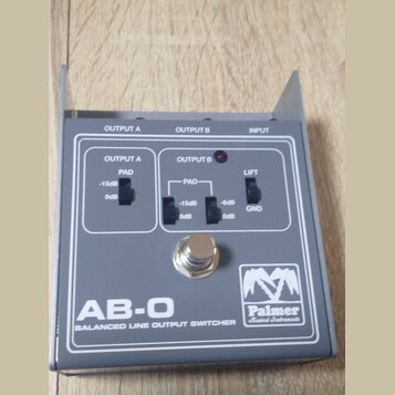 Šedý kovový přístroj s nápisem AB-O BALANCED LINE OUTPUT SWITCHER a logem Palmer Musical Instruments. Na horní straně jsou tři přepínače s popisky PA…