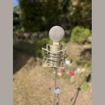 Kondenzátorový mikrofon zlaté barvy s mřížkovou hlavicí, umístěný v drátěném shockmountu na stativu. Na těle mikrofonu je čitelně napsán nápis MERCUR…