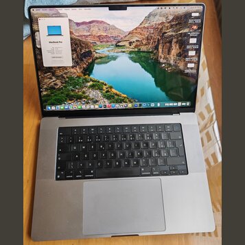 Stříbrný notebook MacBook Pro s otevřeným displejem. Na obrazovce je vidět okno „O tomto Macu“ s nápisem MacBook Pro a v pozadí krajinná fotografie.…