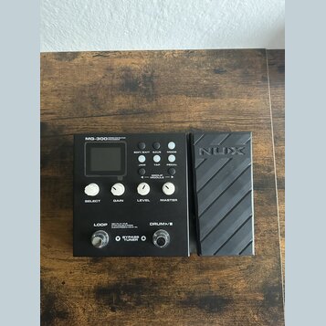 Černý kytarový procesor NUX MG-300 s připojeným expresním pedálem. Na zařízení jsou viditelné nápisy MG-300 MODELLING GUITAR PROCESSOR, EDIT/EXIT, SA…