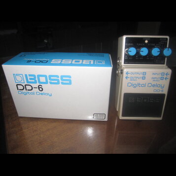 Bílý efekt pedál Digital Delay DD-6 značky BOSS s modrými ovládacími knoflíky. Vedle pedálu je umístěna jeho původní bílá krabice s modrým nápisem BO…