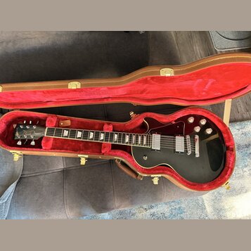 Černá elektrická kytara s tvarem single-cut v hnědém kufru s červeným vnitřkem. Na hlavě je bílé logo Gibson, na hradle jsou dva humbuckery a čtyři o…