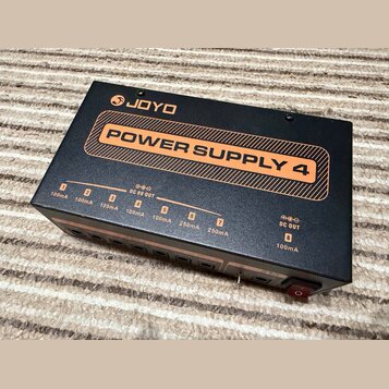 Černý obdélníkový zdroj napájení s oranžovými detaily. Na horní straně je bílé logo JOYO a nápis POWER SUPPLY 4 v oranžovém rámu. Přední strana obsah…