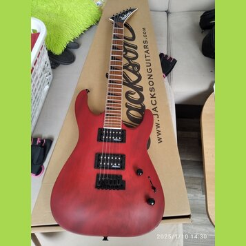 Červená elektrická kytara s tmavým hnedým hmatníkem, položená na kartónové krabici s nápisem „Jackson“ a adresou „WWW.JACKSONGUITARS.COM“. Na hlavě n…