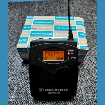 Černý bezdrátový vysílač s displejem, na kterém je text 517.875MHz Y LUDEK01 a indikátor baterie. Na spodní části je bílý nápis SENNHEISER ew 100 G3.…