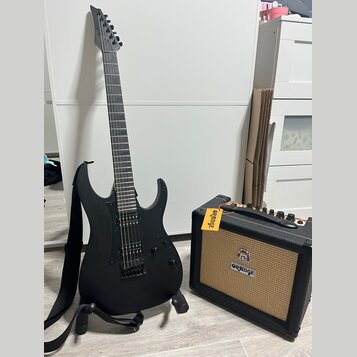Černá elektrická kytara s matným povrchem a černým popruhkem, umístěná na stojanu. Na hlavě kytary je viditelné logo "Ibanez". Vedle kytary stojí mal…