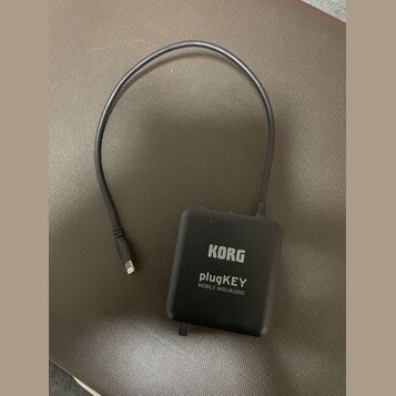 Černý obdélníkový adaptér s připojeným kabelem a konektorem. Na povrchu je bílý nápis KORG, plugKEY a MOBILE MIDI/AUDIO. Povrch zařízení je matný a v…