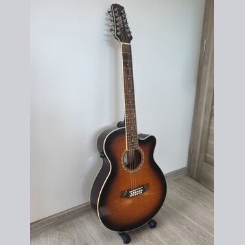 Akustická kytara s cutawayem v tmavě hnědém sunburst provedení. Na hlavě je viditelné logo "Cort". Nástroj je umístěn na černém stojanu u bílé stěny.