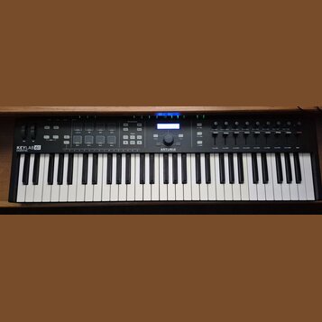 Černý MIDI kontrolér s klaviaturaí. Na levé straně je nápis KEYLAB 61 ESSENTIAL a uprostřed logo ARTURIA. Horní panel obsahuje podsvícené tlačítka, f…