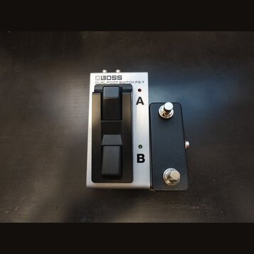 Stříbrný dvoupedálový spínací pedal s nápisem BOSS DUAL FOOT SWITCH FS-7. Vedle něj je umístěn malý černý box se dvěma kovovými přepínači. Na stříbrn…