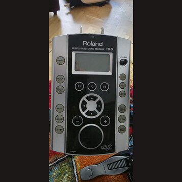 Stříbrný perkusní modul značky Roland s černým předním panelem. Na horní části je nápis PERCUSSION SOUND MODULE TD-9 a v dolní části logo V-Drums. Mo…