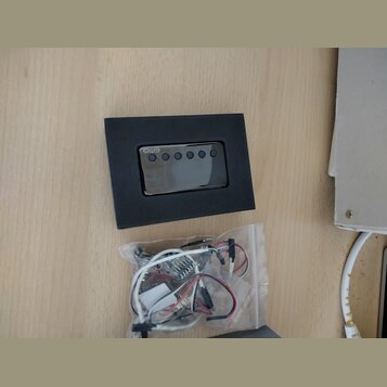 Černý obdélníkový modul s nápisem EMG a pěti kulatými ovládacími prvky. Pod modulem je průhledný plastový sáček s elektroinstalací, kabely a drobnými…