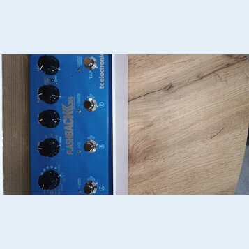 Modrý kytarový efekt s bílým rámem. Na horní straně je nápis FLASHBACK DELAY AND LOOPER a logo tc electronic. Zařízení má čtyři černé otočné knoflíky…