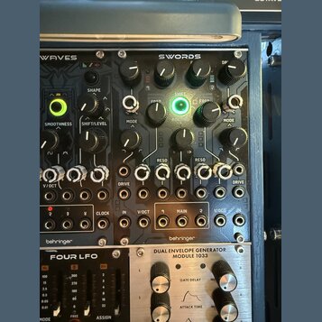 Detailní záběr na rackové moduly syntezátorů. Horní modul je tmavě šedý s nápisy WAVES a SWORDS, značkou behringer v dolní části a řadou knoflíků, př…